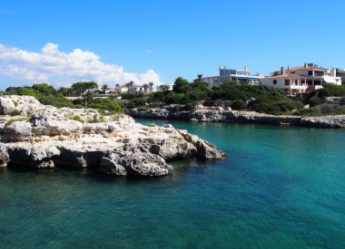sulara bakan kayalıklar ve beyaz evler ile çevrili parlak güneş ışığı ile Menorca 'daki Cala Santandria 'nın doğal manzarası