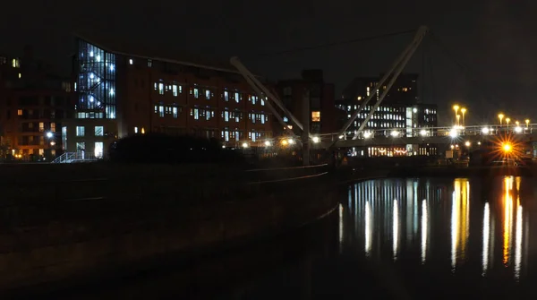 A cityscape view of the canal entrance to the clarence dock area of leeds with a pedestrian bridge crossing the water with reflections of lights and buildings against a night sky with clouds — Stock Photo, Image
