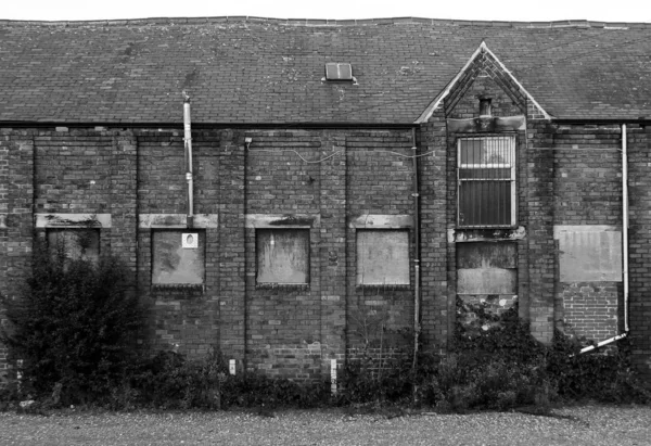 An old abandoned brick factory building with boarded up windows and crumbling decaying walls overgrown with weeds — Stock Photo, Image