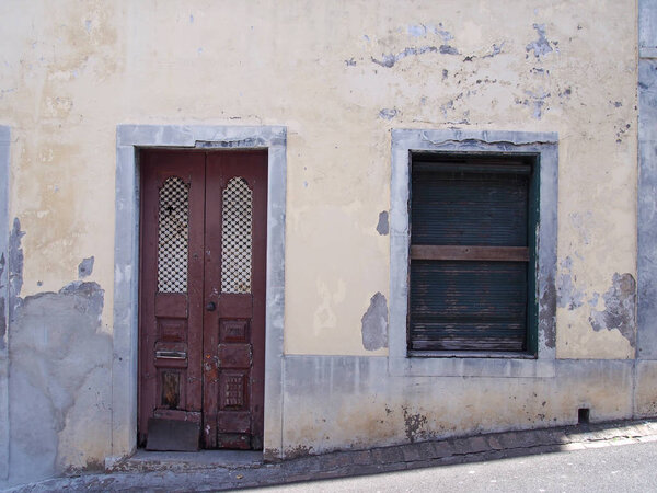 a run down empty abandoned house with crumbling yellow painted walls barred closed shutters and a broken wooden door on a sloping street in funchal portugal