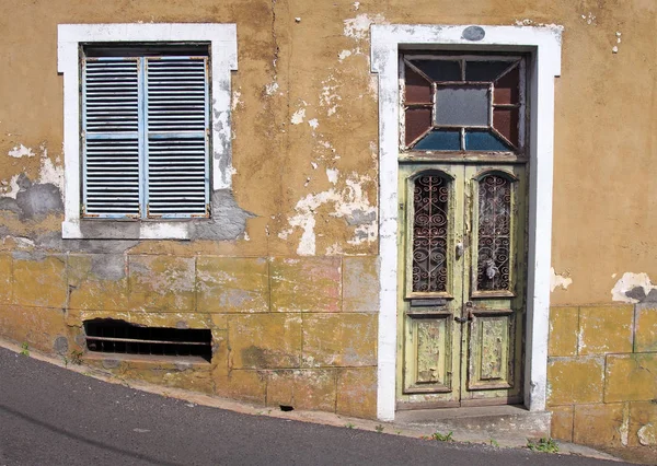 Die Front eines alten verlassenen gelben Hauses mit blauen Fensterläden und verschlossener grüner Holztür mit abblätternder Farbe auf einer abschüssigen Straße — Stockfoto