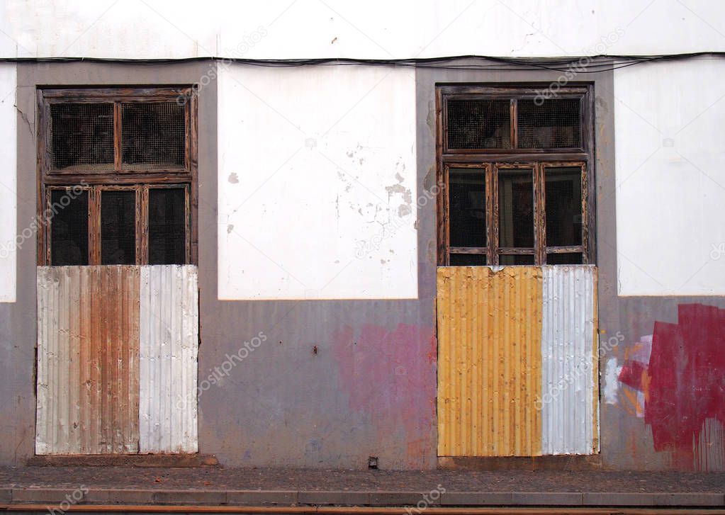 the front of a white and grey painted abandoned house with broken doors and windows boarded up with rusty corrugated iron with splashed of red and yellow