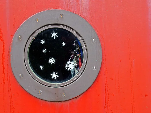 Un primo piano di un oblò rotondo di metallo su una barca rossa con fiocchi di neve e brillantini sulla finestra e fascino lucido appeso all'interno — Foto Stock