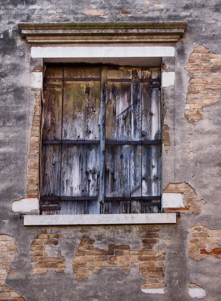Old wooden shuttered window with faded peeling black paint in an ancient brick wall with peeling broken grey cement or plaster