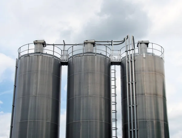 Un gruppo di tre alti serbatoi di stoccaggio chimico in acciaio con tubi di collegamento e scale contro un cielo nuvoloso blu — Foto Stock