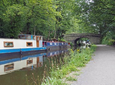 narrow boats and barges moored on the rochdale canal in hebden bridge bext to an old stone footbridge surrounded by green summer trees clipart