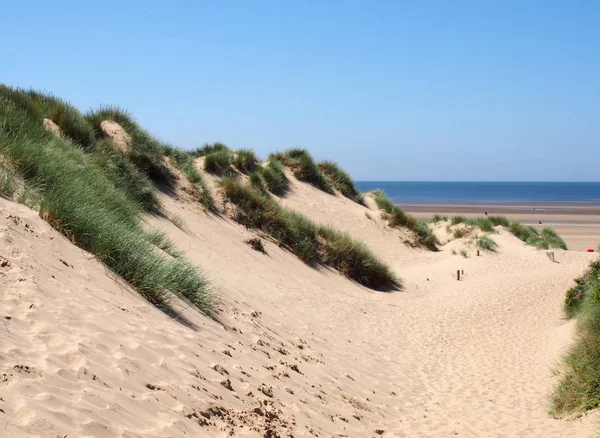 A path through tall grass covered sand dunes leading to a beach and bright blue sea in summer sunlight on the sefton coast in merseyside — Stock Photo, Image