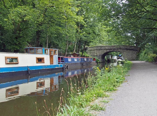 Barche strette e chiatte ormeggiate sul canale di Rochdale nel ponte di Ebden bext per un vecchio ponte pedonale in pietra circondato da alberi estivi verdi — Foto Stock