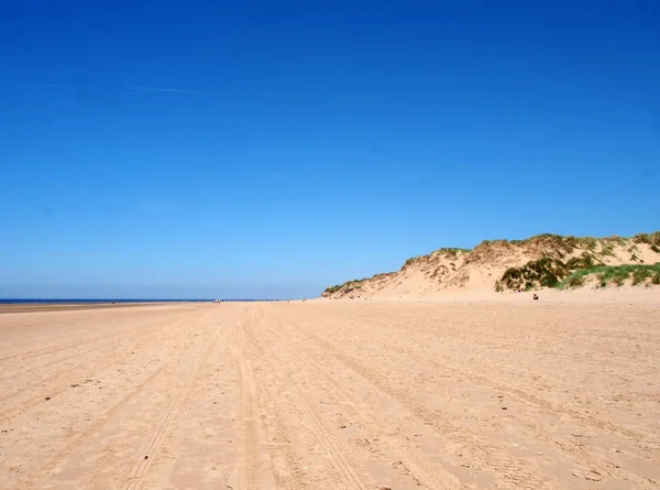 The wide sandy beach on the sefton coast near southport with grass covered sand dunes and unidentifiable people in the distance with a blue summer sky — Stock Photo, Image