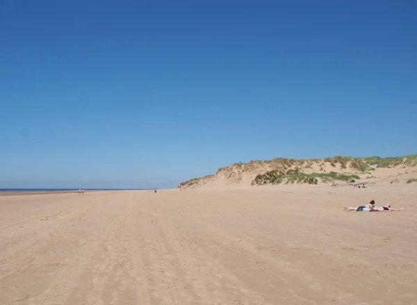 青い空に人々が日光浴や草で覆われた砂丘を望む明るい夏の日に形成された長い砂浜の景色 — ストック写真