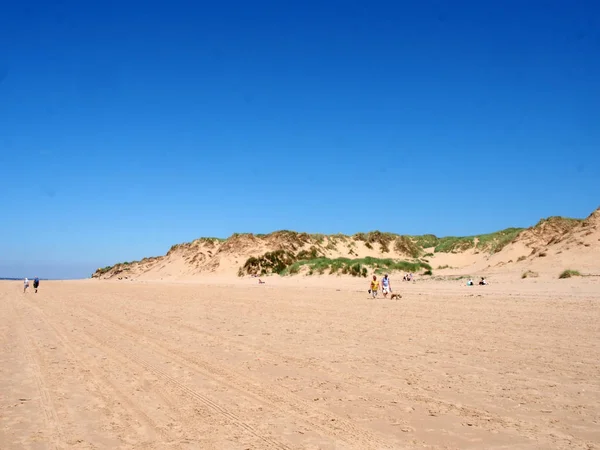 Pessoas caminhando na praia com grama no topo de dunas de areia altas na costa de sefton em merseyside com mar de verão azul e céu — Fotografia de Stock