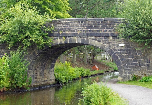 An old stone bridge crossing the rochdale canal at mytholmroyd west yorkshire with a waterside pathway surrounded by trees and bushes — Stock Photo, Image