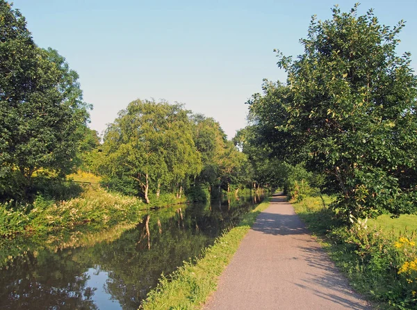 a long straight path along a canal in summer with trees reflected in the water in bright summer sunlight near luddenden in west yorkshire