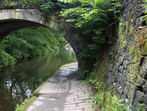 Um caminho ao longo da travessia do canal de rochdale sob uma ponte de pedra velha coberto com vegetação com árvores e um barco estreito na distância — Fotografia de Stock