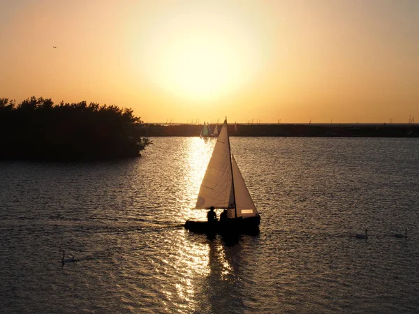 Ein Boot, das auf einem See über den Pfad eines goldenen Sonnenuntergangs segelt, mit nicht identifizierbaren Menschen in Silhouette und Schwänen, die im ruhigen Wasser schwimmen — Stockfoto