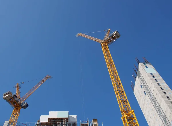 Mavi gökyüzüne karşı büyük beton kulesi olan büyük bir inşaat alanında çalışan iki uzun sarı vinç. — Stok fotoğraf
