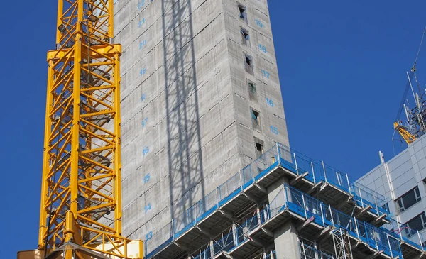 Close-up van een grote stedelijke bouwplaats met een gele torenkraan die een schaduw werpt op een groot betonnen gebouw en veiligheidshekken tegen een blauwe lucht — Stockfoto