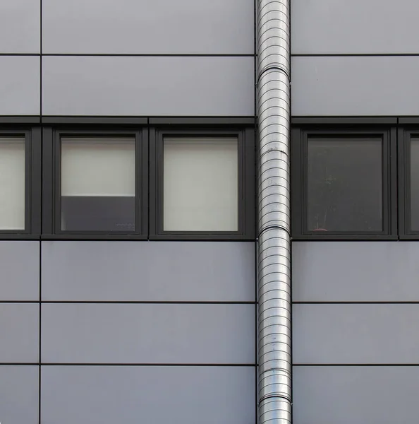 grey cladding panels on an industrial building with a row of back window frames and a silver pipe running up the wall