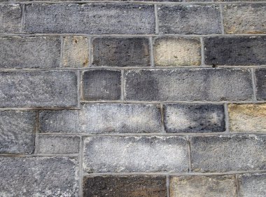 Büyük, düzensiz kumtaşı bloklarından yapılmış büyük, eski, siyah bir taş duvarın tam kare görüntüsü.