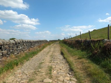 Batı Yorkshire kırsalında taştan duvarlarla çevrili uzun dar bir kır şeridi manzarası çimlerle kaplı çayırlar ve güneşli mavi bir gökyüzü