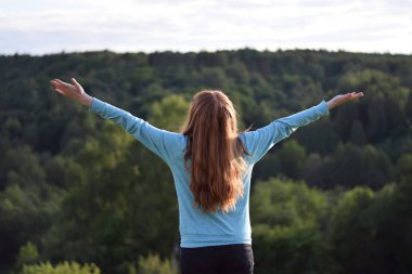 Kızıl saçlı Genç kız eller yayılan bir mavi kazaklı. Arkadan görünüm. Peyzaj dağlar orman arka plan. Özgürlük seyahat ilham kavramı