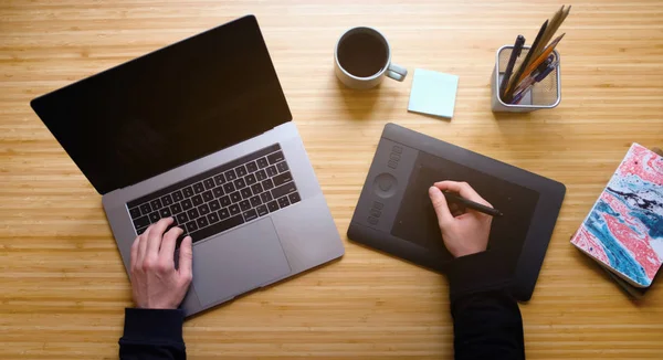 Creative Professional Graphic Designer Illustrator Workspace. Hands Drawing. Workplace Aerial Top View. Modern Wooden Table Desk with Laptop, Graphic Tablet, Stylus, Coffee Mug, Notepad, Lamp Flat Lay