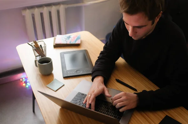 Young Handsome Graphic Designer Illustrator Man at Work. Black Sweatshirt. Cozy Workplace Wooden Table, Laptop, Tablet Pen, Coffee Mug, Sketchbook. Creative Design Working Process. Dark Atmosphere.