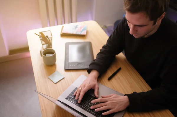 Young Handsome Graphic Designer Illustrator Man at Work. Black Sweatshirt. Cozy Workplace Wooden Table, Laptop, Tablet Pen, Coffee Mug, Sketchbook. Creative Design Working Process. Sunny Atmosphere.