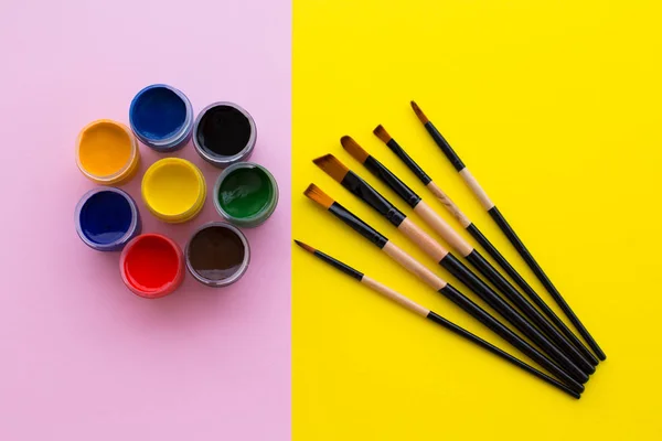 在粉红色背景上精美排列的原色罐 以及在黄色背景上绘制的一组画笔 — 图库照片