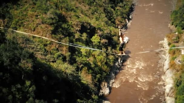 Fiatal férfi gyaloglás a slackline kötélen között Rocks magasan a föld felett, zöld erdő és áramló folyó a háttérben — Stock videók