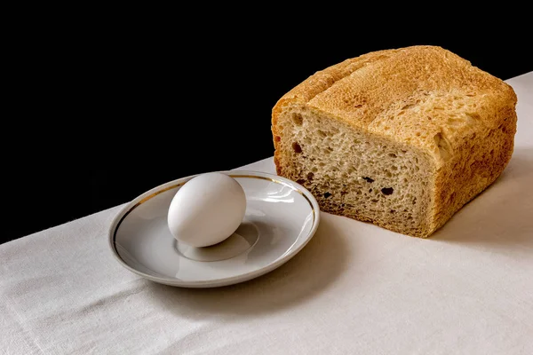 Still life, classics of the genre, chicken egg, saucer, bread and white cloth on a black background.