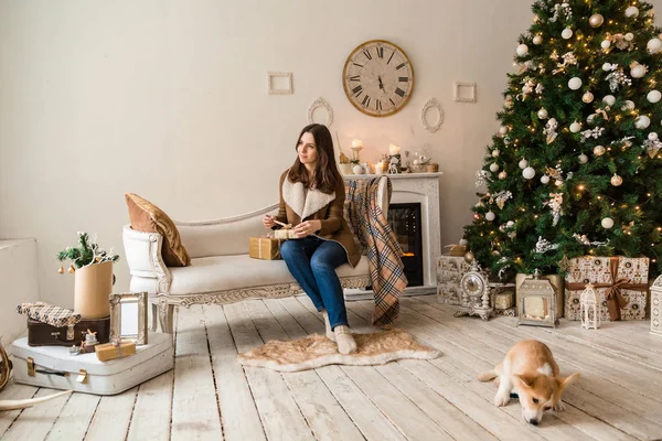 Ein Mädchen in einer warmen Jacke sitzt vor dem Sofa und spielt mit einem Welpen walisischen Corgi-Strickjacke, über Geschenke nachdenkend — Stockfoto