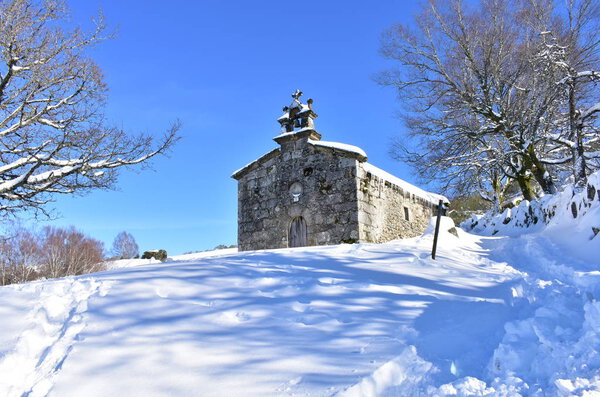 Piornedo, Lugo, Spain. San Lorenzo Hermitage with snow, ice and icicles. Sunny day, trees and blue sky.