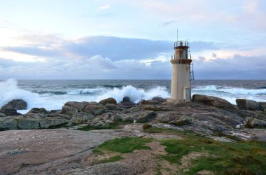 Eski deniz feneri ve dalgalar gün batımı ışığı ile kayalara karşı kırma. Muxia, İspanya.