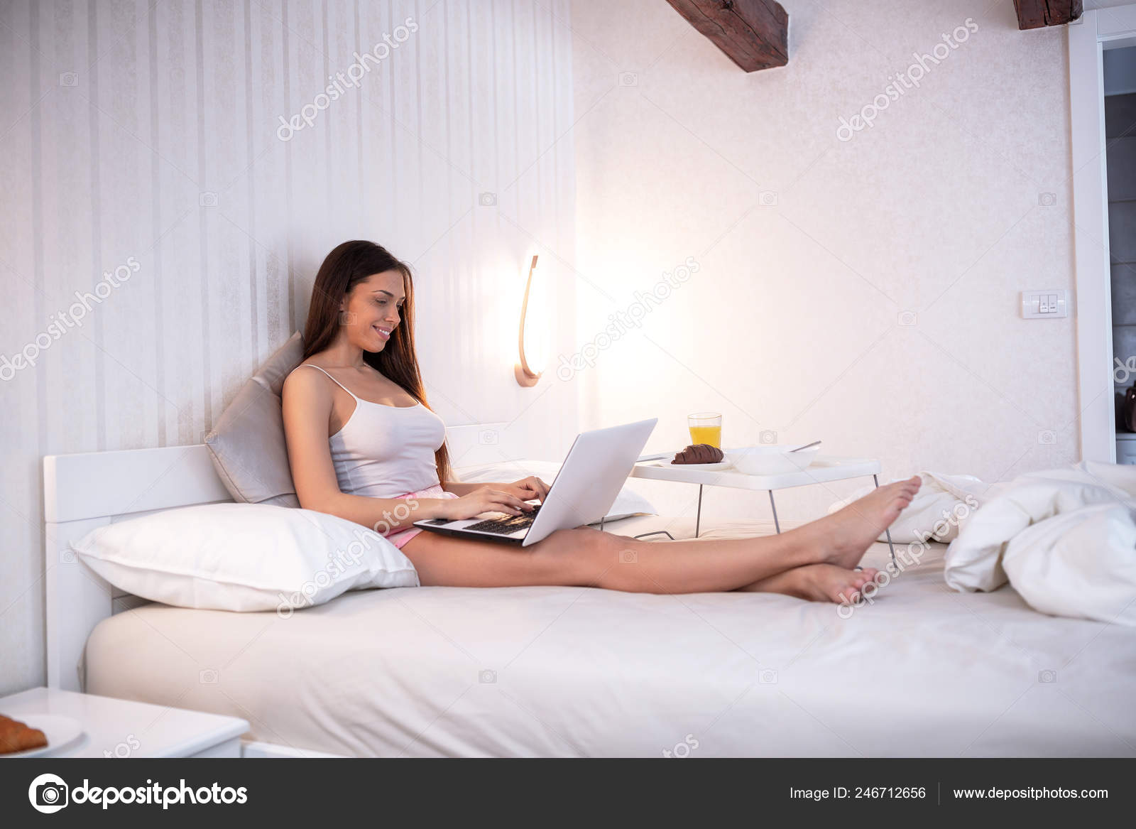 Lat morgon med hennes laptop, unga vackra kvinnan sitter på sängen 
