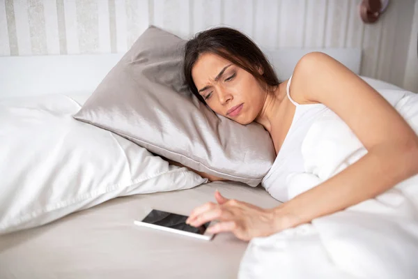 Tired beautiful young woman in bed holding her mobile phone