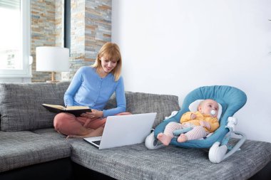 Genç anne ve bebeği kanepede, evde çalışıyorlar.