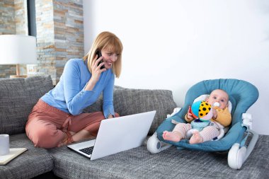 Annesi ve küçük bebeği kanepede, kadın telefonda konuşuyor.