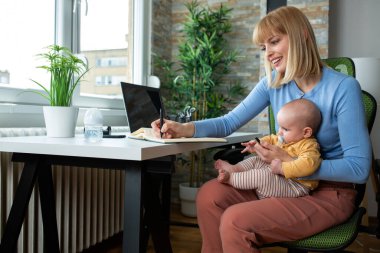 Sevgi dolu anne çalışırken not alıyor ve aynı zamanda kucağında bir bebek tutuyor.