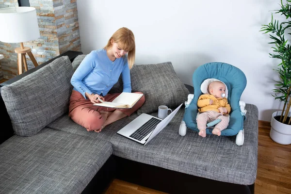 Genç anne ve bebeği kanepede, kadın not tutuyor.