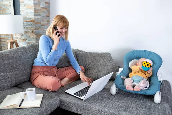 Annesi ve küçük bebeği kanepede, kadın telefonda konuşuyor.