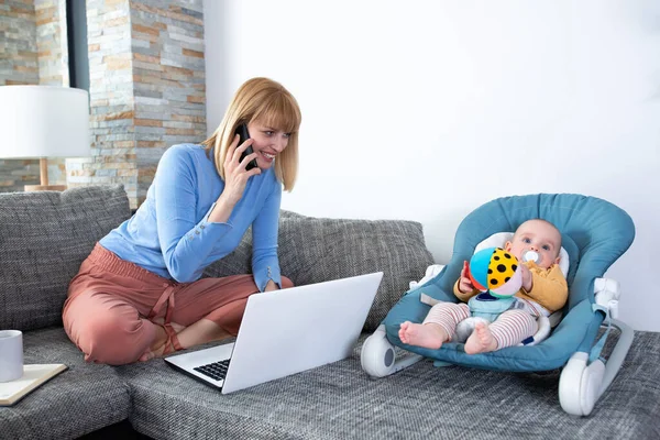 Annesi ve küçük bebeği kanepede, kadın telefonda konuşuyor.