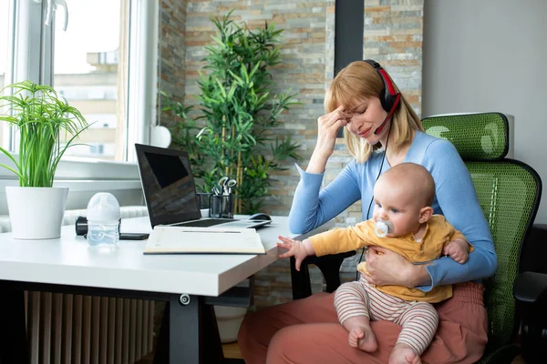 若い母親が座って赤ちゃんを抱えて仕事をしたり仕事をしたり頭を悩ませたり — ストック写真