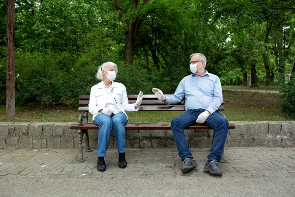 Senior people practicing social distancing and wearing face masks and gloves as they sit on the park bench