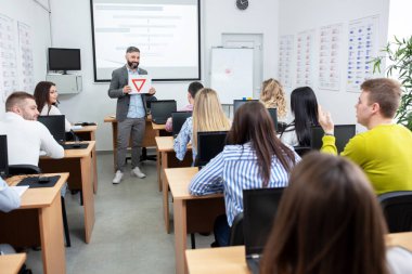 Sürücü eğitmeni öğrencilerine kağıda çizilmiş trafik işaretlerini gösteriyor ve bunun anlamını soruyor. 