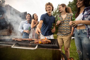 Genç adam, arkadaşlarıyla eğlenirken eti barbeküye koyuyor.