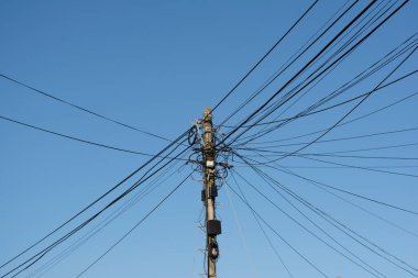 Elektrik ve telekomünikasyon kablolarının büyük karışıklığı mavi gökyüzü arka planının altındaki tahta bir direğe bağlı.