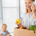 Улыбающаяся мать держит продукты во время разговора на смартфоне с малышом в детском кресле на заднем плане