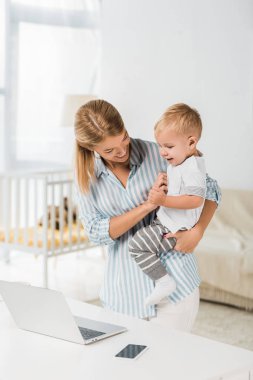 Danışma laptop ve smartphone ile yakın duran ve gülümseyen yürümeye başlayan çocuk elinde tutan neşeli anne 