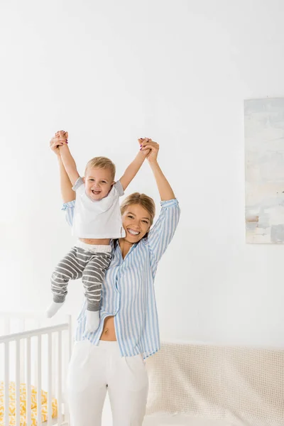 Alegre Madre Mirando Cámara Sosteniendo Sonriente Niño Las Manos Habitación —  Fotos de Stock